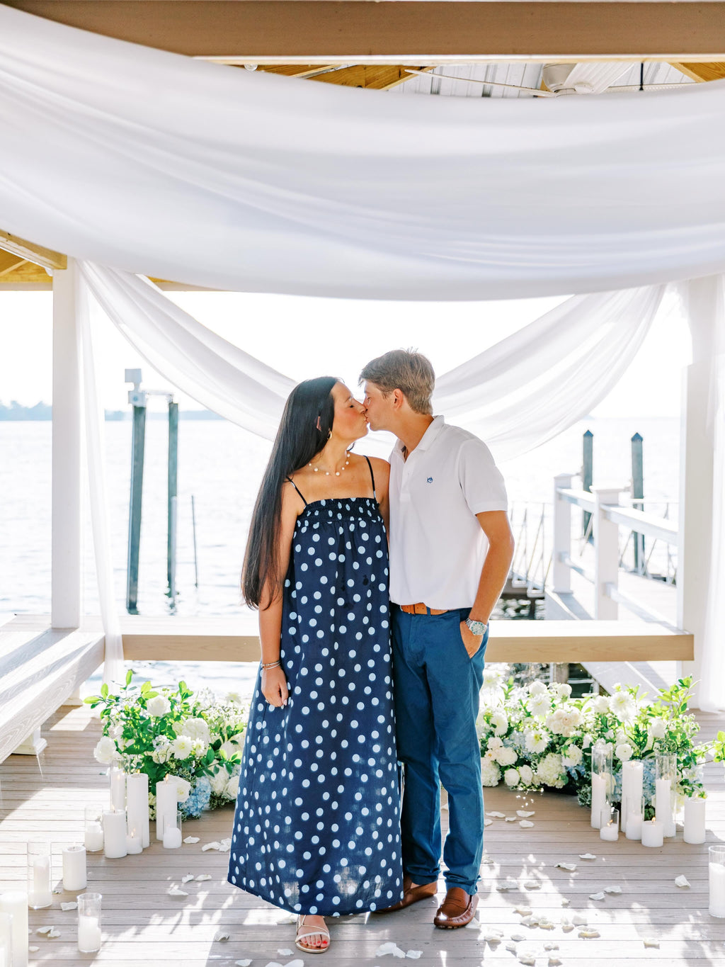 A couple embracing and kissing on a wooden dock decorated with white draped fabric, candles, and floral arrangements. The woman wears a navy blue strapless maxi dress with white polka dots, and the man wears a white polo shirt and blue jeans.