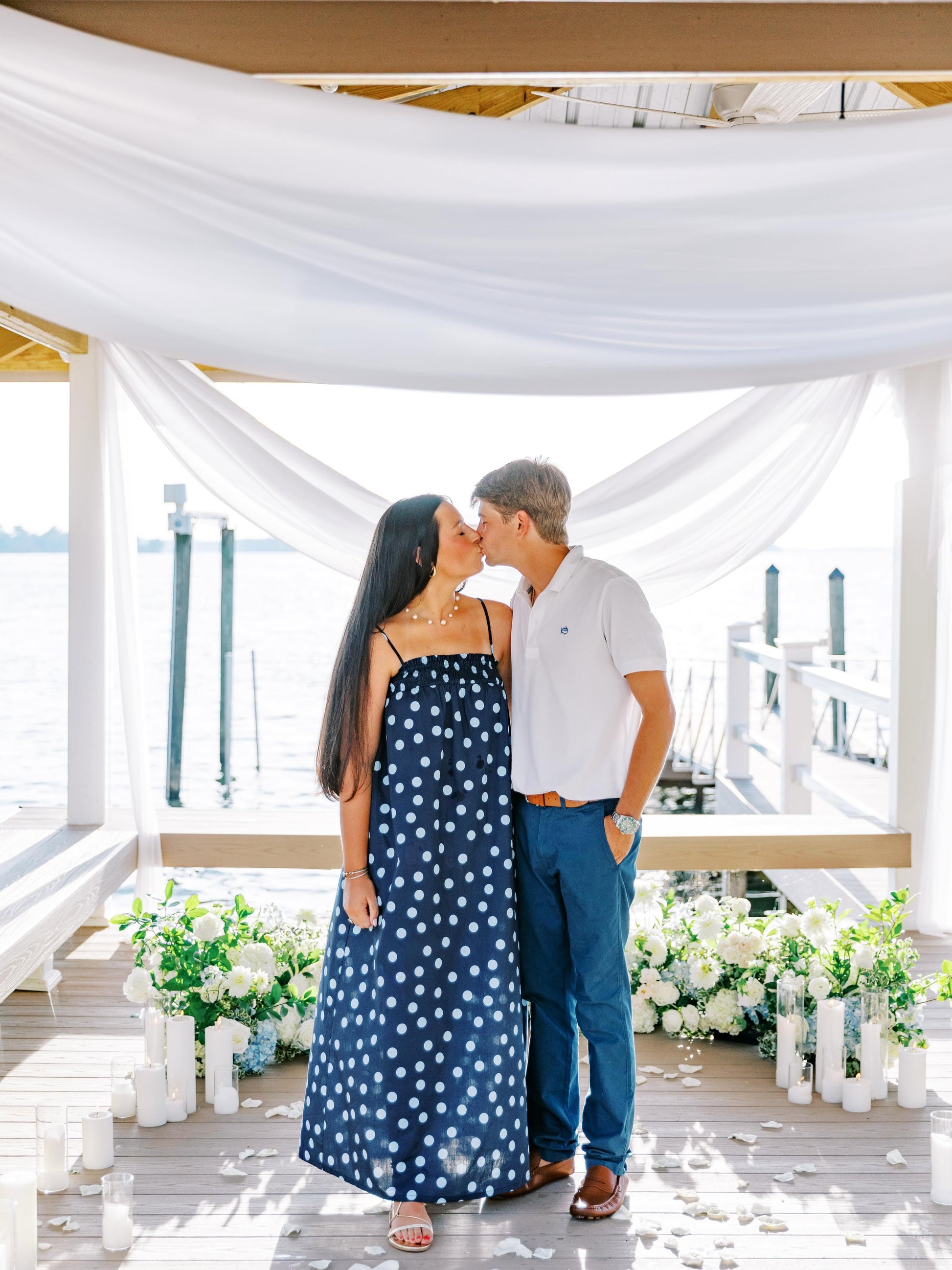 A couple embracing and kissing on a wooden dock decorated with white draped fabric, candles, and floral arrangements. The woman wears a navy blue strapless maxi dress with white polka dots, and the man wears a white polo shirt and blue jeans.