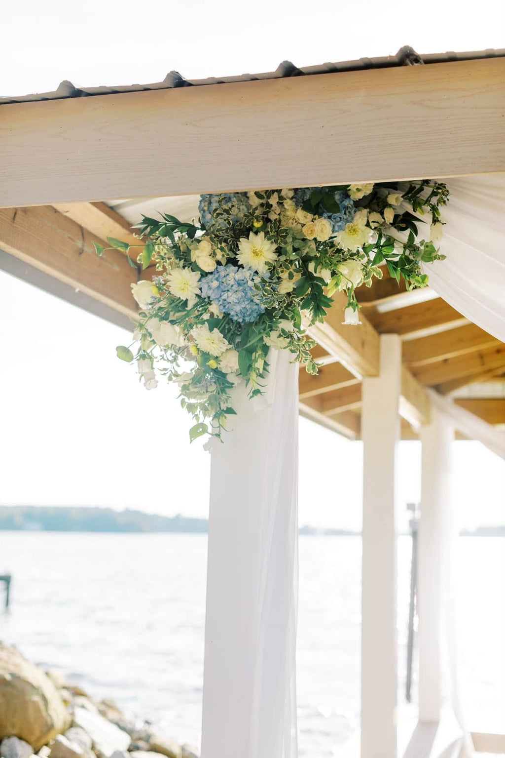 A floral arrangement of white and light blue flowers with green foliage hanging from a wooden structure above white draped columns, with water and rocks visible in the background.