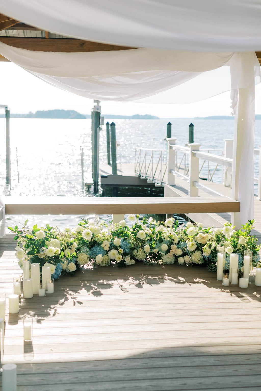 A beautifully decorated outdoor wedding dock or porch setup with a floral arch of white and light blue flowers, white pillar candles, and draped white fabric overhead, overlooking a body of water.
