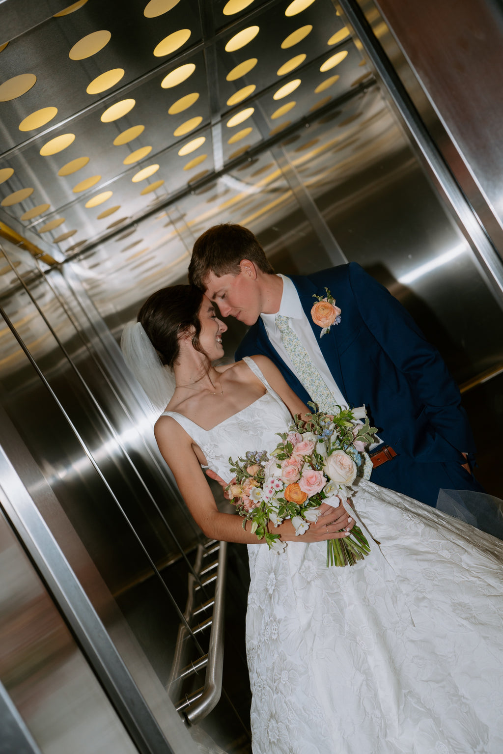 A bride in a white sleeveless wedding gown holding a pastel flower bouquet leans in to kiss her groom inside a metallic elevator. The groom wears a navy blue suit with a white shirt and a peach boutonniere.