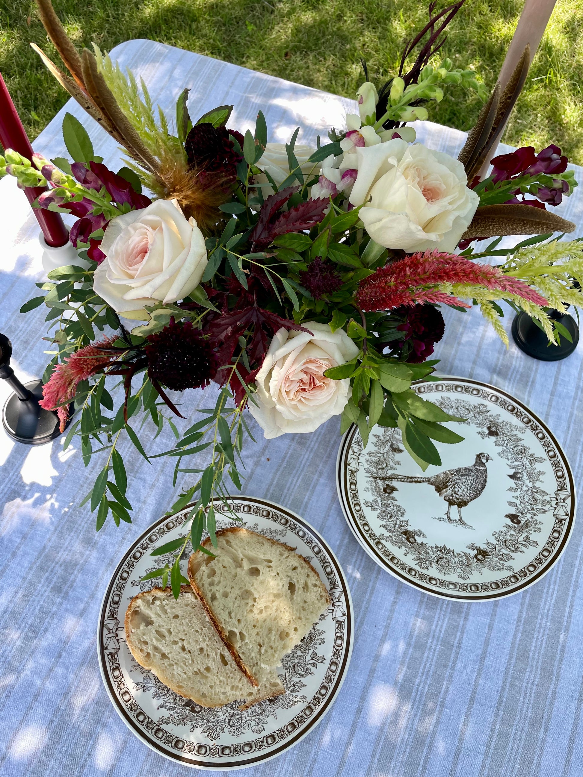 Floral arrangement with white roses and greenery on a tablecloth, accompanied by decorative plates and a sandwich.