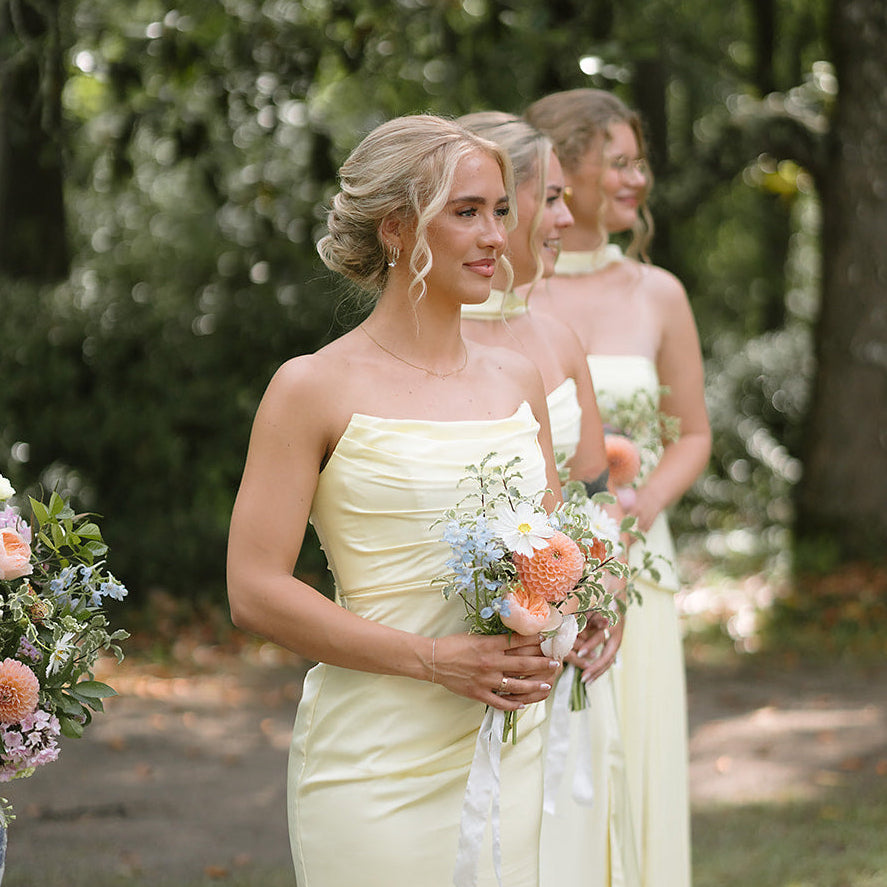 A woman in a pale yellow strapless gown stands outdoors holding a bouquet, with two other women in similar dresses behind her in a wooded setting.