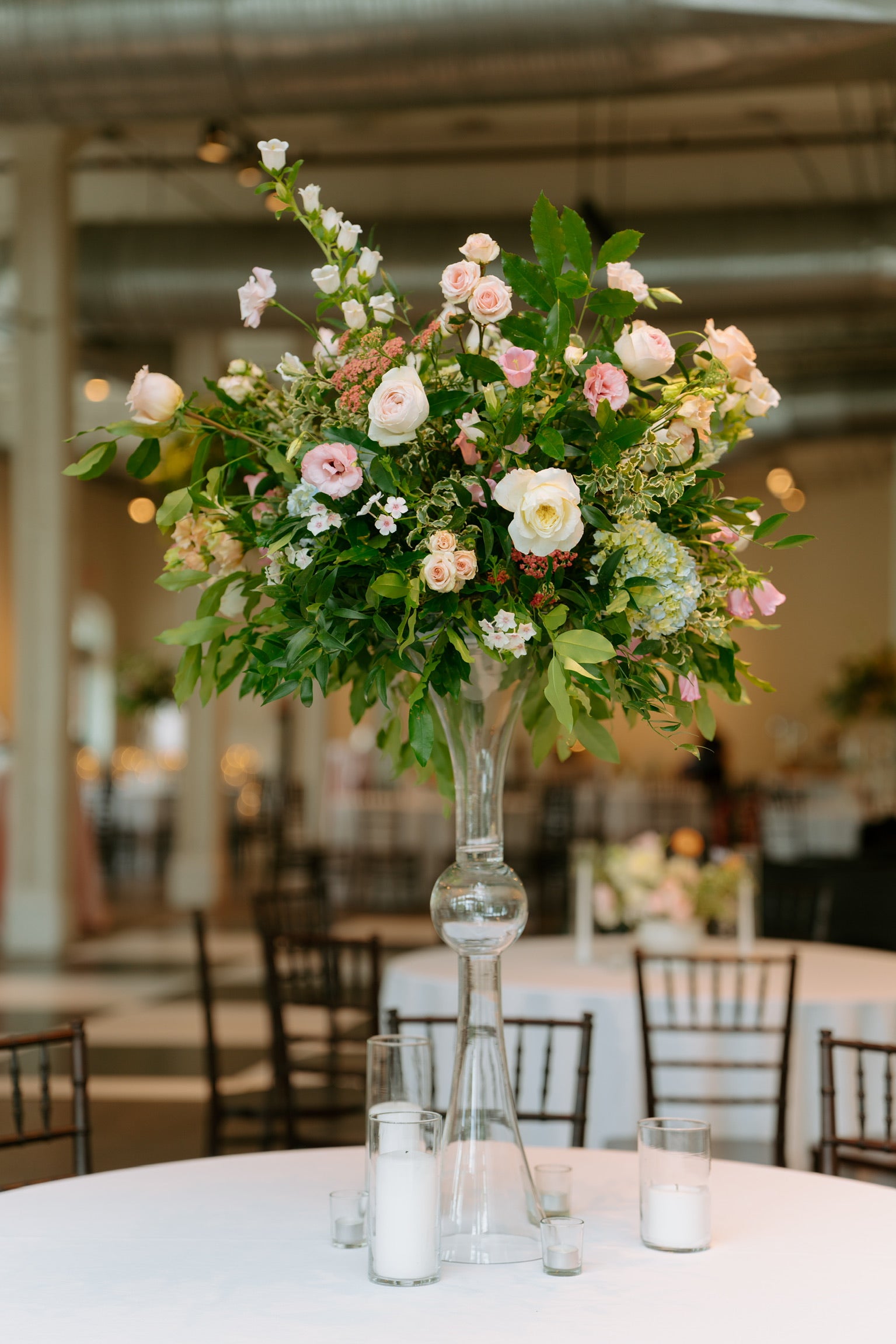A large floral centerpiece with pink and white flowers and green foliage arranged in a tall clear glass vase on a white table, with blurred event tables and chairs in the background.