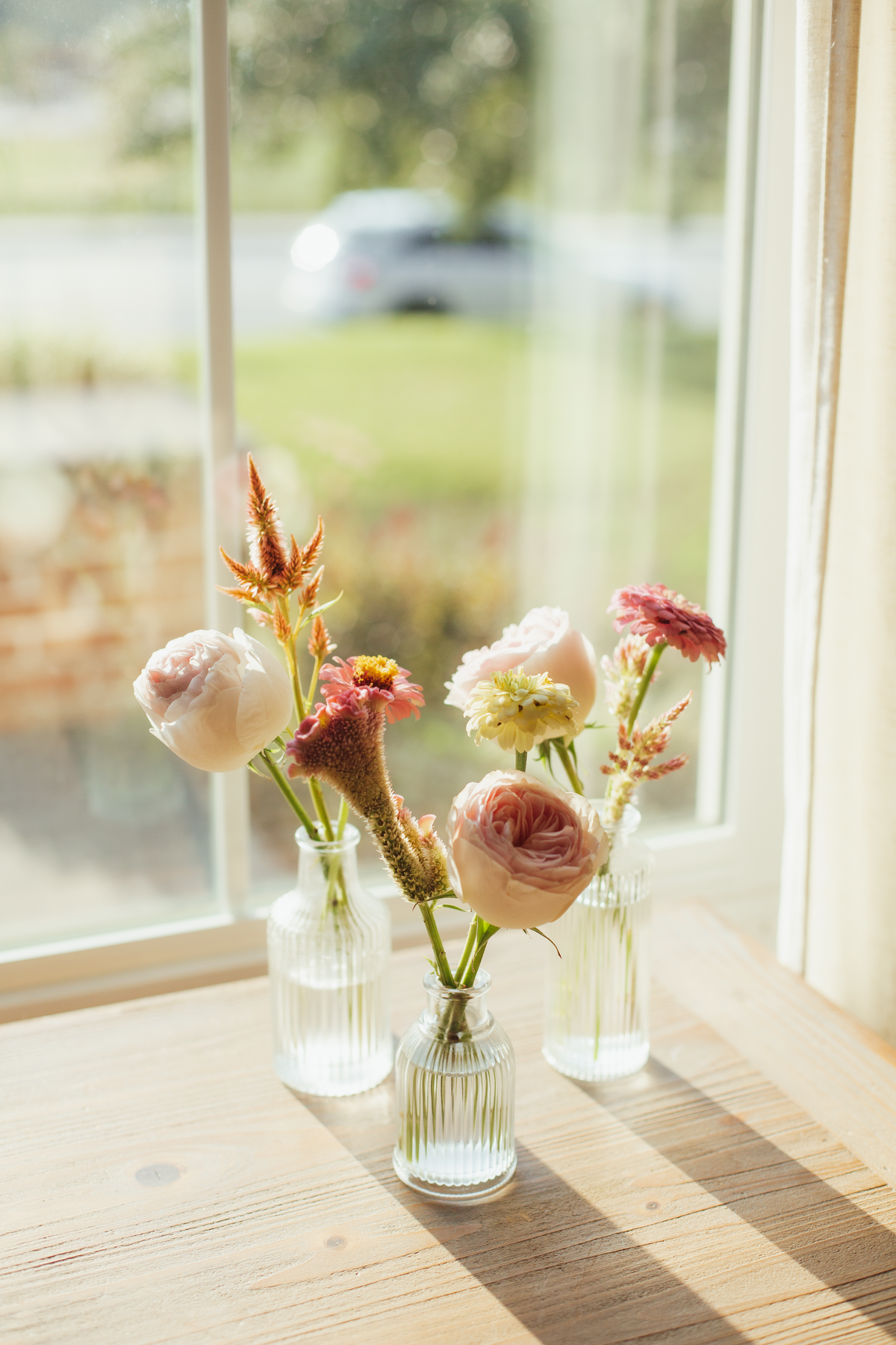 Three small glass vases with flowers on a wooden surface near a window.
