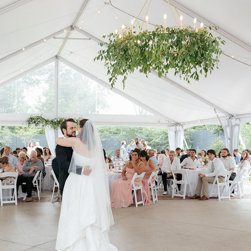 A bride in a white wedding gown and veil dances under a large white event tent, with guests seated at round tables around her. The tent has string lights, floral arrangements, and greenery hanging from the ceiling.