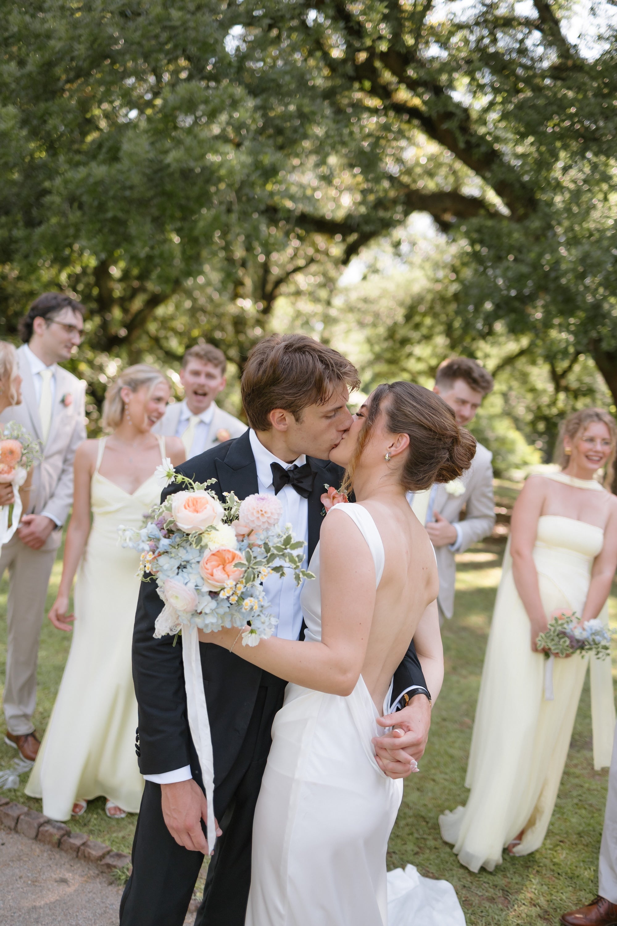A bride in a white sleeveless wedding gown and a groom in a black tuxedo kiss in an outdoor park setting, surrounded by smiling bridesmaids and groomsmen holding bouquets.